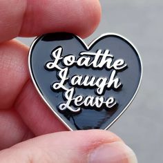 someone holding a black heart shaped pin with the words loathe laugh leave on it