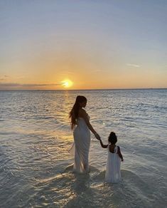 a woman holding the hand of a child in the ocean at sunset with the sun setting behind them