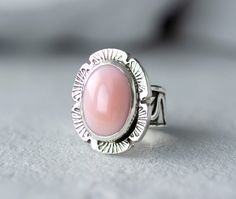 Bold but dainty Pink Conch Silver ring on a unique silver band that has been handmade using scrap 925 Sterling Silver. The band has been fused, oxidized then lightly polished. At its widest the band is 3/8" then tapers to 1/8". The oval cabochon is 14mm x 10mm and is in a bezel setting accented with stamped, cut out square wire. Add a touch of Coastal Cowgirl attitude to your outfit with our simple, but elegant Pink Conch Silver Statement Ring. Whether you're shopping for yourself or are searchi Silver Pink Opal Ring Gift, Silver Pink Opal Ring For Gift, Pink Opal Rings As A Gift, Oval Pink Opal Jewelry For Gifts, Oval Pink Opal Jewelry Gift, Pink Rings With Polished Finish For Gift, Handmade Sterling Silver Pink Rings, Handmade Pink Sterling Silver Rings, Artisan Pink Ring As Gift