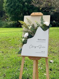 an easel with flowers and greenery on it sitting in the middle of a field