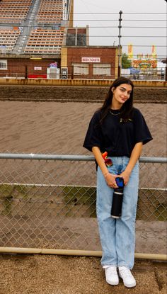 a woman standing next to a fence holding a skateboard in her hand and looking at the camera