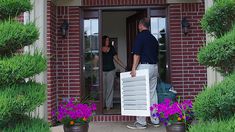 a man and woman carrying boxes into the front door of a house with potted plants