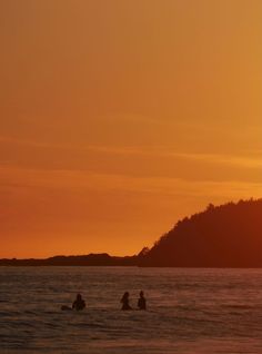 three people in the water at sunset with a hill in the background and an orange sky