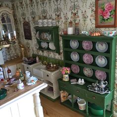 a doll house kitchen with green cabinets and plates on the shelves, dishes in front of it