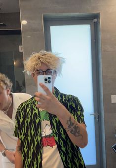 a man with blonde hair taking a selfie in front of a mirror while wearing a green and black shirt