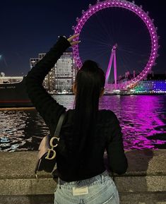 a woman standing next to the water with a ferris wheel in the background at night