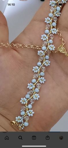 a hand holding a gold chain bracelet with white beads and blue flowers on the side