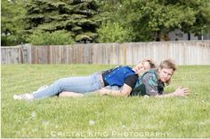 a man and woman laying on the grass in front of a fence with trees behind them