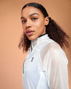 a woman with curly hair wearing a white jacket and posing for the camera in front of a brown background