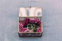 two wedding rings sitting in a glass box filled with purple and pink wildflowers