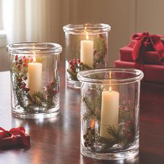 three clear glass candle holders with christmas wreaths and pine branches in them on a table