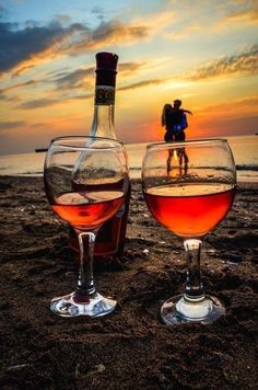 two wine glasses sitting on top of a sandy beach next to the ocean at sunset