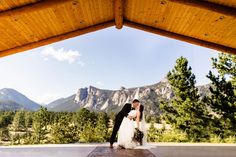 a bride and groom standing under a wooden structure with mountains in the backgroud