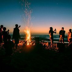 a group of people standing around a fire on top of a beach next to the ocean