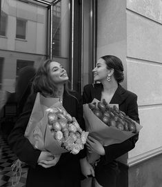 two women are holding flowers and smiling at each other while standing on the sidewalk in front of a building