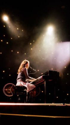 a woman sitting at a piano in front of spotlights on a stage with lights behind her