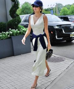 a woman in white dress and blue hat walking down the street with her hand on her hip