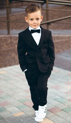 a young boy in a tuxedo and bow tie standing on the sidewalk with his hands in his pockets