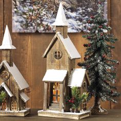 two small wooden houses with snow on the roof and trees next to them, sitting on a table in front of a wood paneled wall