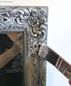 a person holding a paintbrush in front of a silver frame with an ornate design on it