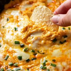 a hand dipping a tortilla chip into a casserole dish with cheese and green onions