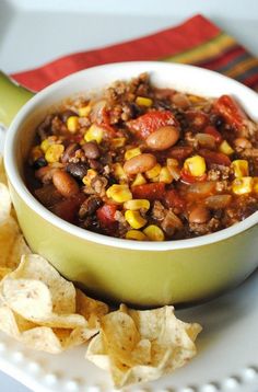 a green bowl filled with chili and corn on top of a white plate next to tortilla chips