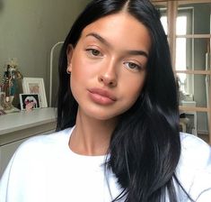 a woman with long black hair wearing a white shirt and gold hoop earrings looking at the camera