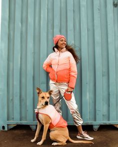 a woman in pink jacket standing next to a brown and white dog wearing silver pants