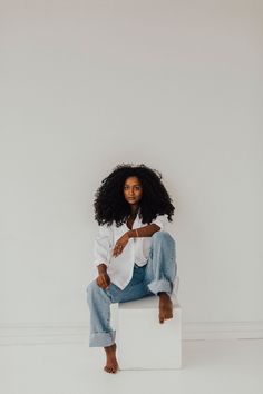 a woman sitting on top of a white block wearing jeans and a white button up shirt