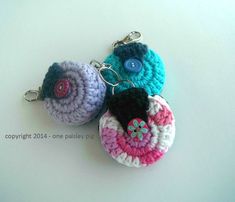 three crocheted keychains are sitting on a white surface, one has a blue button and the other has a pink flower