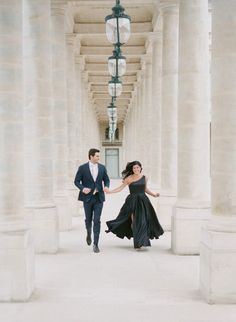 a man and woman in formal wear walking down an empty hallway with columns on either side