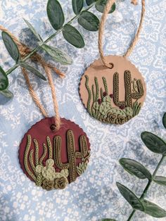 two ceramic ornaments with cactus designs hanging from twine on a blue and white tablecloth