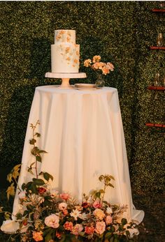 a wedding cake sitting on top of a table covered in flowers and greenery next to a wall