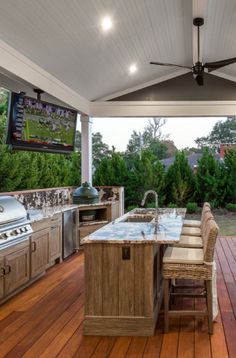 an outdoor kitchen with grill and television on the wall, surrounded by wood flooring