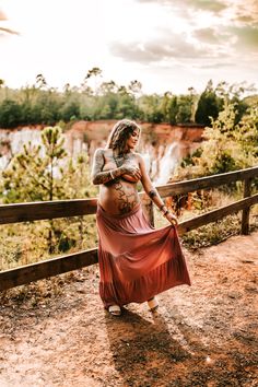 a pregnant woman in a long red dress is standing on a wooden fence looking at the water