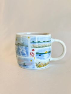 a coffee cup with pictures of the ocean and mountains on it's side, sitting on a white surface