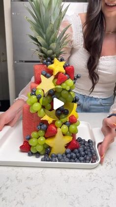 a woman cutting up a fruit decorated cake on top of a white plate with grapes, pineapples and blueberries