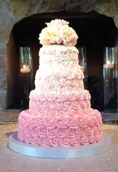 a pink and white wedding cake with roses on it sitting in front of a fireplace