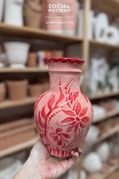 a hand holding a red and white vase in front of shelves with pottery on it