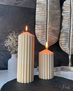 two white candles sitting next to each other on top of a black table with silver vases behind them