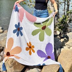 a woman standing on rocks holding up a colorful flowered crocheted blanket with tassels