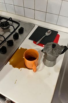 a stove top with a cup of liquid on it next to a tea pot and spoon