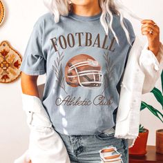 Gear up for game day with our Football Athletic Club Comfort Color Tee. Crafted for comfort and style, this tee is perfect for fans who want to show their support in a laid-back yet spirited way. Emblazoned with the emblem of our Football Athletic Club, this tee exudes team spirit and camaraderie. Whether you're cheering from the stands or watching from home, our tee ensures you'll be representing your team in style. Made from premium materials, this tee offers unparalleled comfort and durabilit Collegiate Graphic Print T-shirt For Game Day, Collegiate Game Day T-shirt With Team Name, Fall Varsity T-shirt With Logo Print, Varsity T-shirt With Team Logo For Football Season, Athletic Heather T-shirt With Team Name For Game Day, Game Day T-shirt With Team Name In Athletic Heather, Game Day Sports Fan T-shirt With Crew Neck, Athletic Heather T-shirt For Game Day, Game Day Football Season T-shirt With Logo