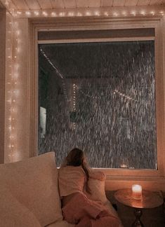 a woman sitting on a couch in front of a window covered with rain falling down