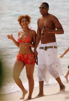 a man and woman walking on the beach in swimsuits, with one holding a cell phone