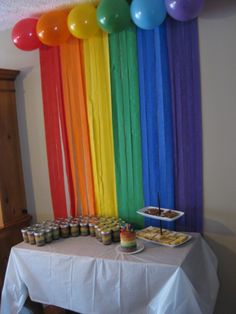 there is a table with food and balloons in the air above it, along with rainbow colored streamers hanging from the ceiling