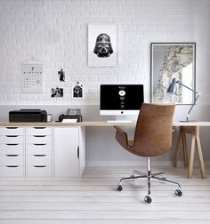 a desk with two computers on it in front of a white brick wall and wooden chairs