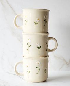 three white cups with daisies painted on them sitting side by side in front of a marble background