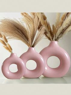 three pink vases sitting next to each other on a white surface with dried grass