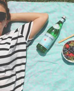 a woman laying on the grass next to a bowl of fruit and a bottle of beer